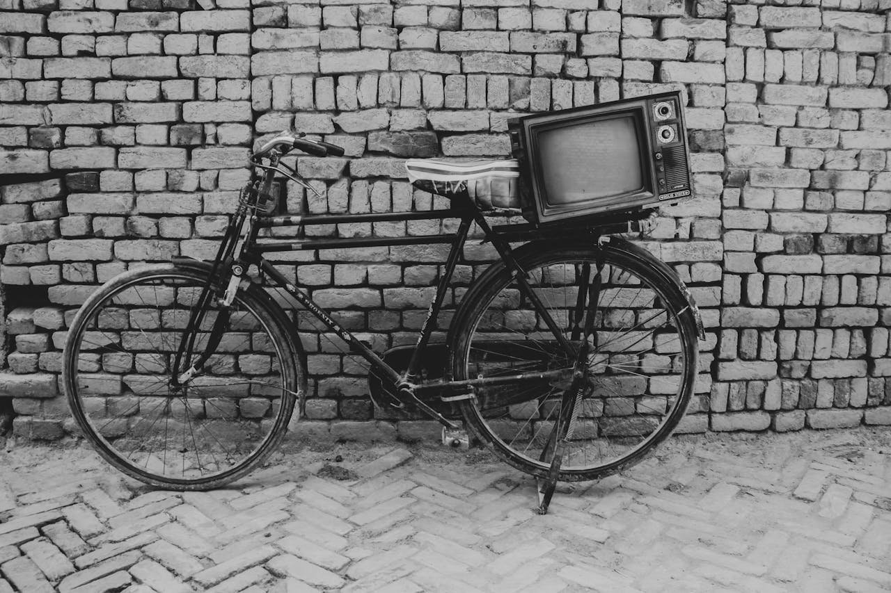A vintage bicycle with an old television set mounted, set against a rustic brick wall in grayscale.
